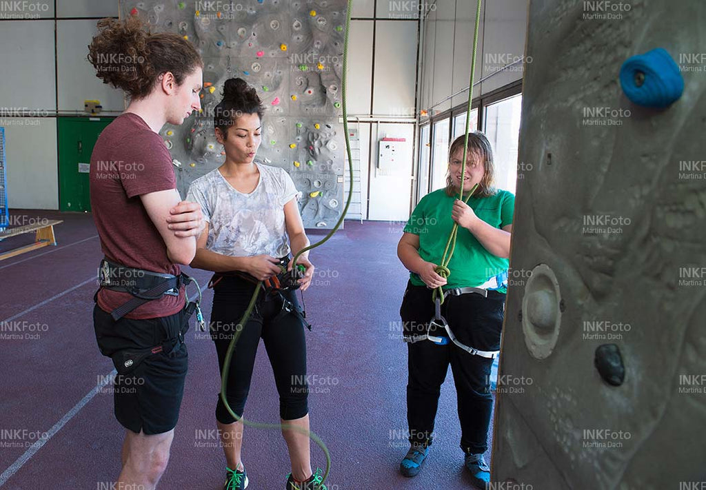 Drei junge Erwachsene vor einer Kletterwand beim Anseilen.