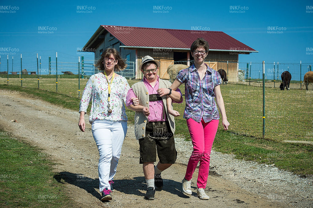 Zwei Frauen und ein Mann wandern Arm in Arm bei Sonnenschein zu einem Bauernhof.