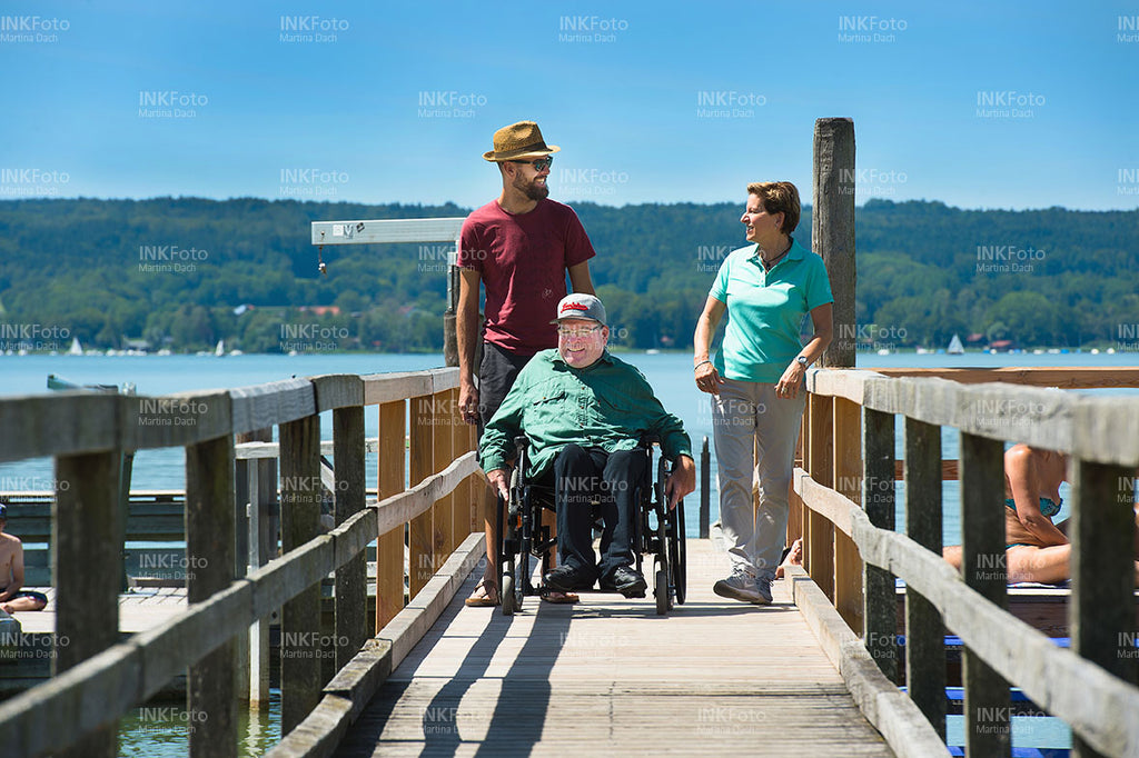 Mann im Rollstuhl am See begleitet von einer Frau und einem Mann mit Sonnenhut