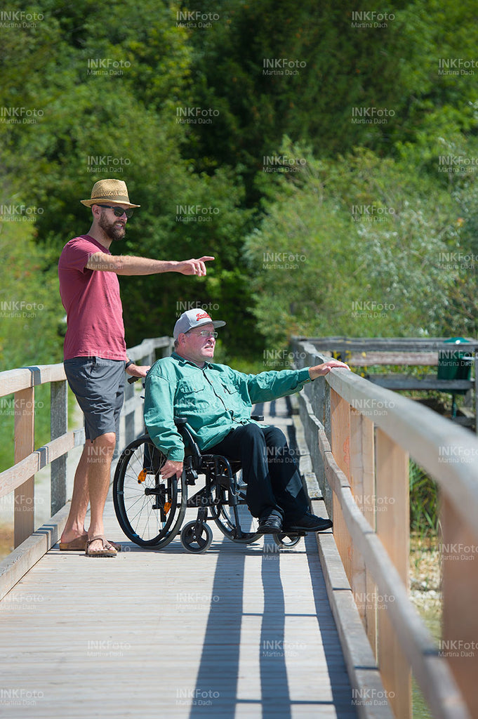 Mann im Rollstuhl am See begleitet von einem Mann mit Sonnenhut