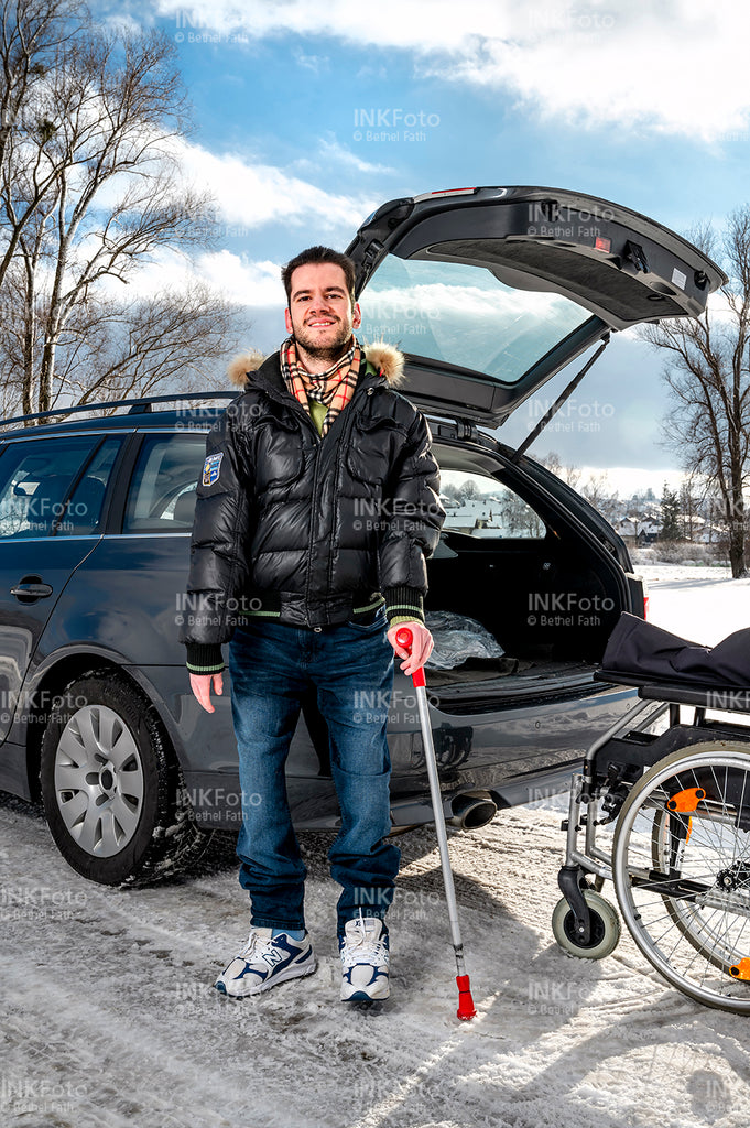 Junger Mann mit Krücke und Rollstuhl vor einem Auto.
