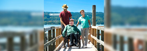 Lächelnder Mann im Rollstuhl auf einem Steg am See im Sommer, begleitet von einem jungen Mann mit Sonnenhut.