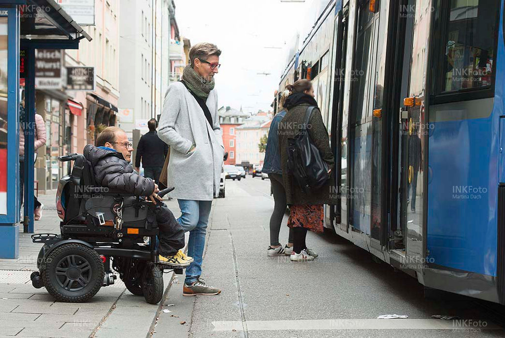 Ehepaar im Rollstuhl an einer Haltestelle mit Straßenbahn in der Stadt