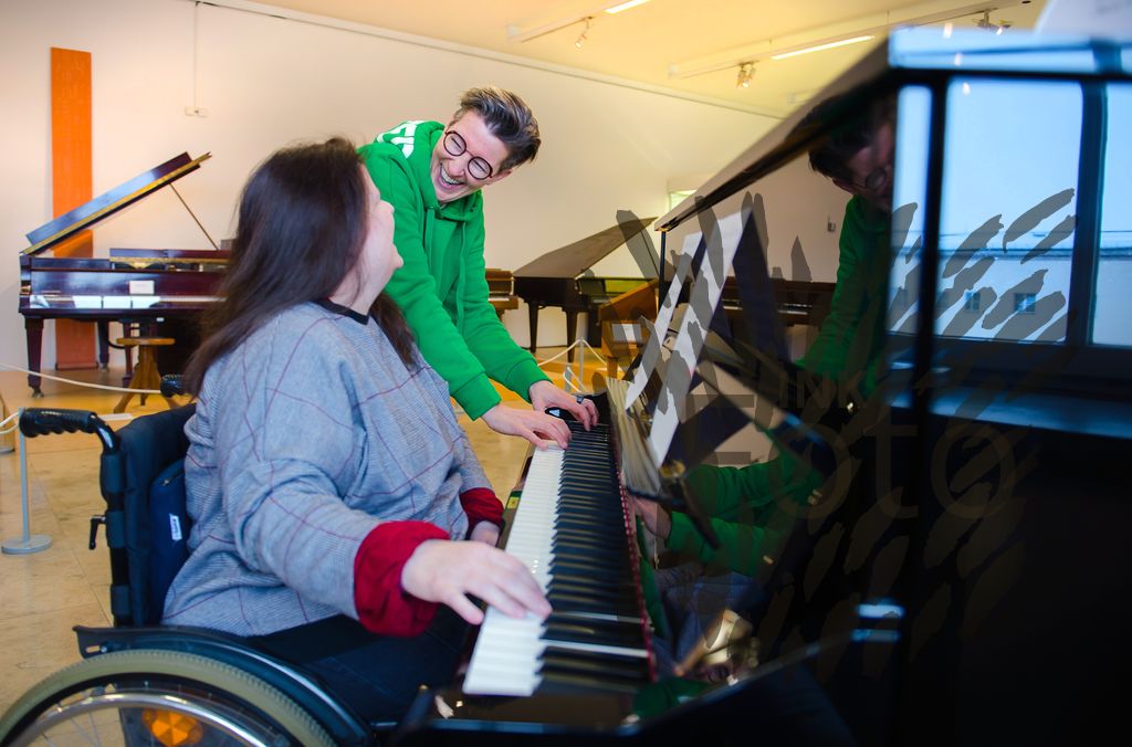 Zwei Frauen vor einem Klavier, eine Frau spielt das Instrument während die andere Frau ihr interessiert über die Schulter schaut.