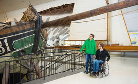 Eine ältere Frau mit Brille und ein junger Mann im Rollstuhl betrachten im Technischen Museum ein ausgestelltes Boot.