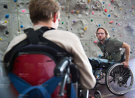Zwei junge Männer im Rollstuhl vor einer Kletterwand in einer Sporthalle.