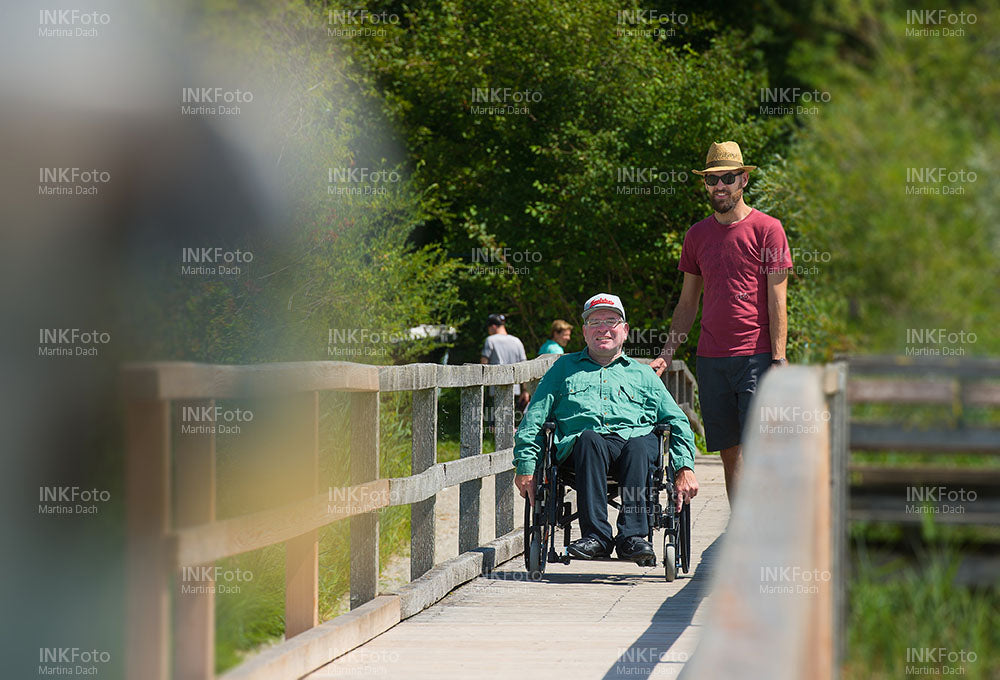 Mann im Rollstuhl am See begleitet von einem Mann mit Sonnenhut