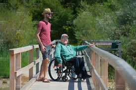 Lächelnder Mann im Rollstuhl auf einem Steg am See im Sommer, begleitet von einem jungen Mann mit Sonnenhut.