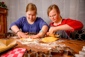 Zwei junge Mädchen sitzen an einem Tisch und rollen Teig für Plätzchen aus.