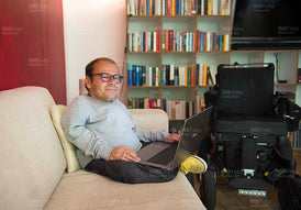 Mann mit Behinderung und Rollstuhl auf der Couch mit Laptop im Homeoffice