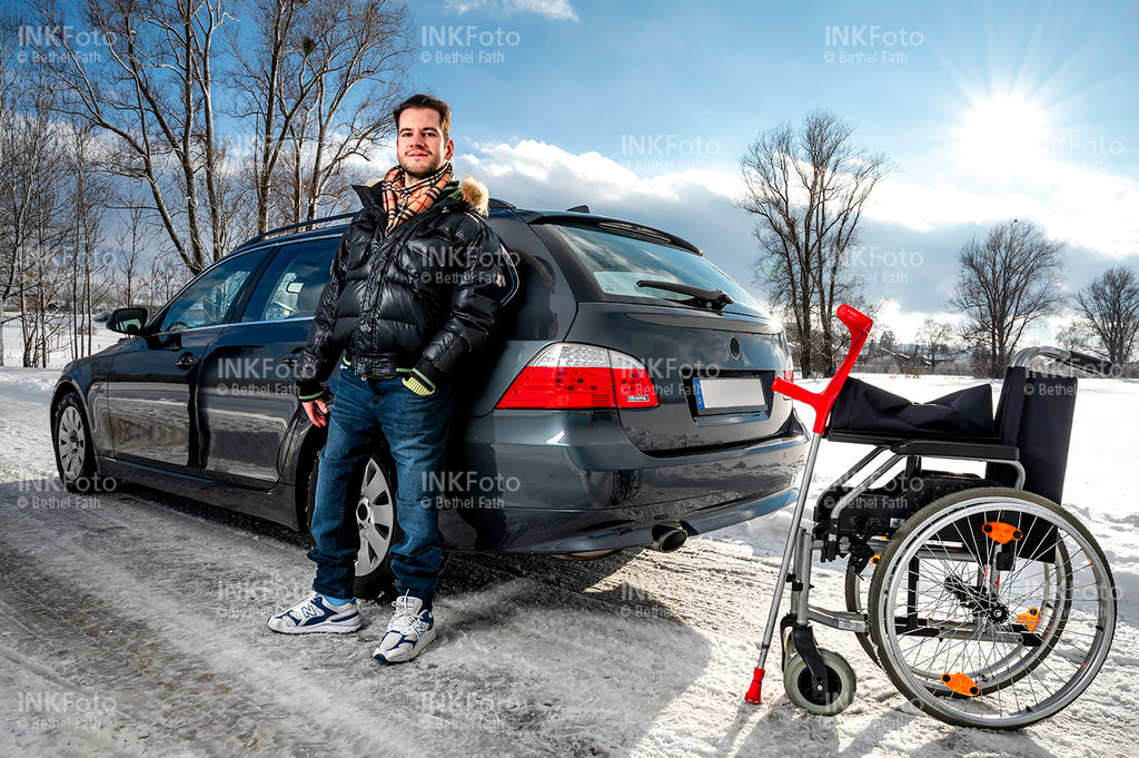 Junger Mann mit Krücke steht lässig vor einem Auto neben ihm der Rollstuhl.