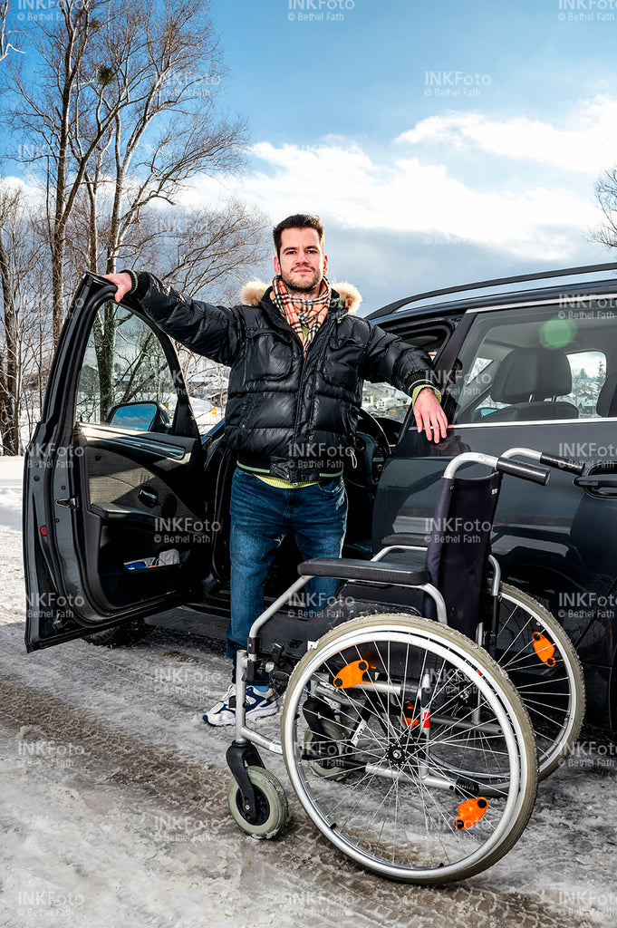 Junger Mann mit Rollstuhl und Krücke vor einem Auto.